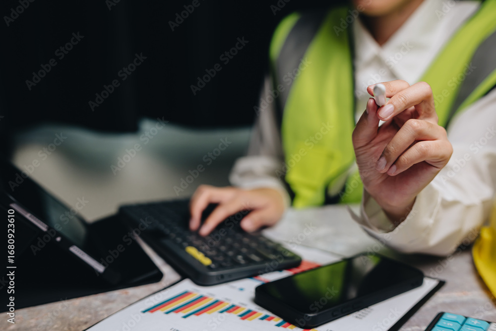 Poster professional in safety vest working on laptop with tablet and smartphone while analyzing data on a d