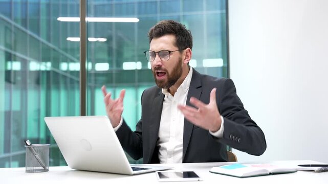 Angry businessman arguing talking on video call using laptop computer sitting at workplace in business office. Excited dissatisfied boss in formal suit scolds subordinate, solving stressful situation.