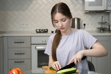 Young girl cutting cucumber on wooden board, preparing a healthy salad with fresh vegetables like...