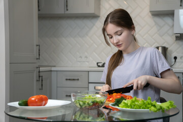 Teen girl skillfully slices a vibrant red bell pepper on a wooden cutting board, preparing a healthy salad in her contemporary kitchen, surrounded by fresh ingredients