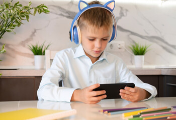Boy wearing headphones with phone in his hands using smartphone technology while doing homework. Concept of children's dependence on mobile games. Online Education.