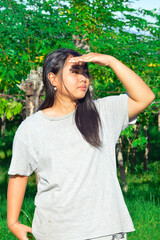 A girl stands under tropical trees, lifting her hand to block sunlight while gazing into the distance, enjoying the peaceful morning countryside air.