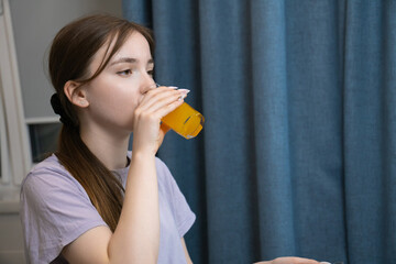 Fototapeta premium Young girl enjoying a glass of orange juice while browsing her smartphone. Casual indoor setting with blue curtains in the background, conveying a relaxed and modern lifestyle