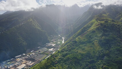 Aerial view of a lush green valley in Tahiti, French Polynesia, with sun rays penetrating through clouds, illuminating a river flowing amidst the mountains and industrial buildings