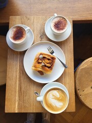 Coffee date with children. 1 cappuccino cup, 1 cinnamon roll, 2 cups of babyccino (milk) are  on wooden rustic table.