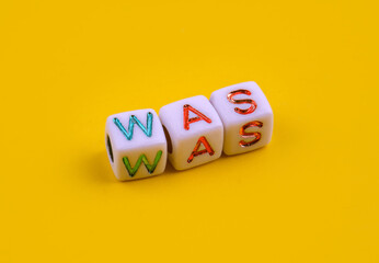 Three white dice with colorful engraved letters spelling the word "WAS" placed on a bright yellow background. The playful cubes symbolize language, creativity, learning and word-based games.