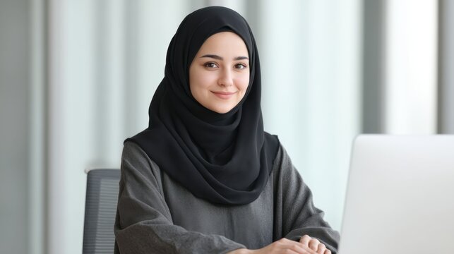 Young woman in black hijab smiling while working on a laptop in a modern office setting with soft natural light and minimalistic decor