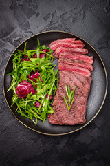 Grilled and sliced Flank beef steak on plate with salad. black background. top view