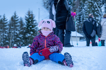 young child, little girl sledding in colorful winter outfit and pink bunny hat sits on snow,...
