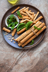 Plate with different breadsticks, basil oil and fresh basil on a brown granite background, vertical shot with space, top view