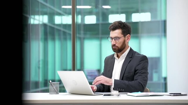 Thoughtful businessman working on laptop sitting at desk at workplace in business office. Manager thinking about problem solving. The worker is engaged in a task or is busy with a project on computer.