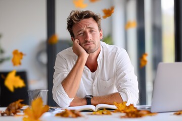 Frustrated man leaning on hand at desk while autumn leaves fall outside the office. Late-season creative block and emotional fatigue in quiet modern workspace.