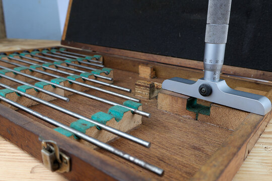 A set of vintage depth micrometers is displayed in an open wooden case on a rustic wooden table. The precise measuring tools with their various extension rods.
