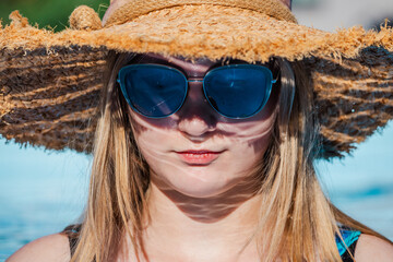 Close-up portrait of a young woman wearing a straw hat and blue sunglasses by the pool, enjoying a...