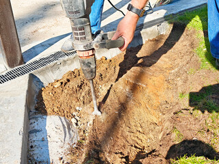 Worker uses drill to excavate soil for construction project in urban setting during daylight hours