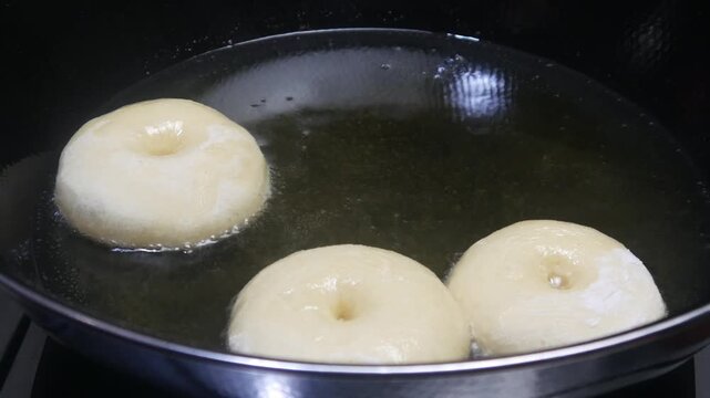 The process of frying donuts in a pan filled with hot oil. Some of the dough is still white and half cooked, while others have begun to turn brown. Making homemade donuts