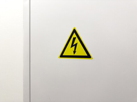 Yellow triangular high voltage sign with black lightning bolt on white electrical cabinet door, safety warning symbol indicating danger of electric shock, industrial risk prevention label