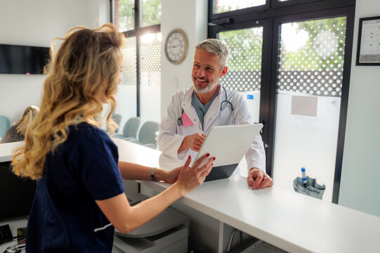 Senior male doctor smiling while showing and explaining information on a tablet to a young female nurse at the hospital reception desk, engaging in a lively discussion about patient care