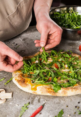 Hands of a chef skillfully adding fresh arugula to a gourmet pizza topped with vegetables, showcasing culinary artistry and vibrant ingredients in a kitchen setting