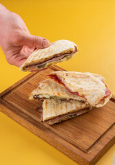 Hand holding delicious sandwich with layers of meat and cheese, placed on wooden board against...