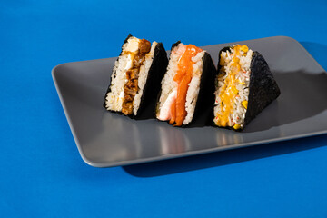 Three colorful sushi onigiri arranged on a gray plate against a vibrant blue background, showcasing delicious fillings and traditional Japanese cuisine aesthetics
