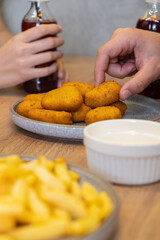 Close-up of hands reaching for golden crispy chicken nuggets on a gray plate, accompanied by dipping sauce and fries, creating a casual dining atmosphere