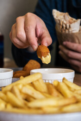 Hand of a person dipping a crispy golden chicken nugget into creamy sauce, with a plate of golden fries and additional dipping sauces in a casual dining setting