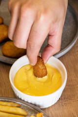 Hand dipping crispy golden nugget into creamy cheese sauce in a white bowl, with fries and additional nuggets on a rustic wooden table, showcasing delicious snack experience