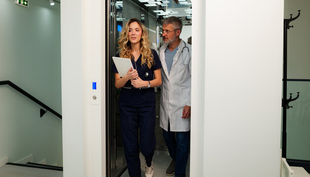 Female nurse holding a digital tablet while senior doctor exiting elevator in hospital hallway, discussing patient medical history and treatment plan, highlighting teamwork in healthcare