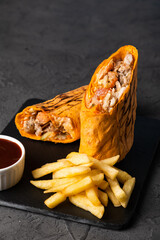 Grilled burrito filled with chicken, vegetables, and spices served with crispy fries and dipping sauce on a black slate plate, showcasing delicious fast food presentation