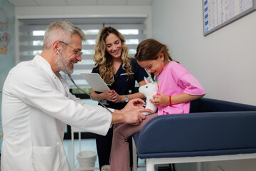 Obraz premium Pediatrician jokingly examining girl's stuffed unicorn with stethoscope while nurse holding digital tablet smiles, creating a playful and reassuring atmosphere during medical checkup