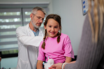 Fototapeta premium Pediatrician examining a little girl with a stethoscope while the mother holds her hand and her plush unicorn, providing comfort and support during the medical checkup