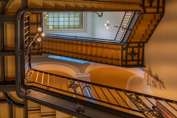 Ornate staircase seen from below, leading to stained glass skylight. Lamps illuminate the elegant...