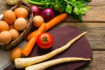 basket of eggs and tomato, spanish onions, carrots, parsley roots, celery, cutting board, old weathered wooden table background