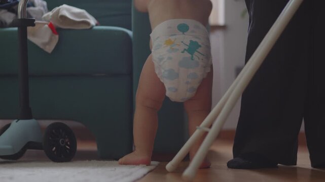 Toddler getting up from floor in diaper near adult legs and baby scooter, exploring living room environment with curiosity and playful mood