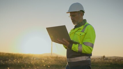Male engineer in helmet and reflective jacket holding laptop in hands. Worker standing in field with wind turbine at sunset. Looking to side. Technician monitoring renewable energy project. - Powered by Adobe