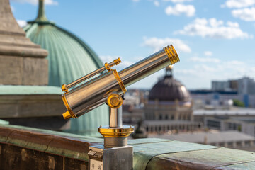 A shiny telescope with golden details stands in front of a green dome. Roofs and the sky are visible in the background.