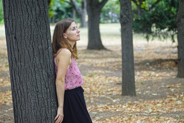 Woman leaning on tree during late summer day in park. Concept of seasonal change, golden leaves,...