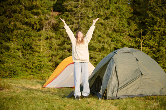 Woman hiker stretching at tent's entrance, warming up in the morning. Young female traveler at the tent looking out at a scenic view. Solo travel, adventure, and enjoying the outdoors. - Powered by Adobe