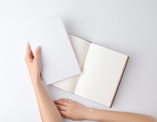 Person holds blank white book over open notebook on a clean white surface, creating a minimalist and serene composition.