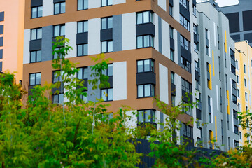A modern residential building with white and brown facades, large windows and balconies. In the foreground are green trees, creating a feeling of coziness and harmony with nature.