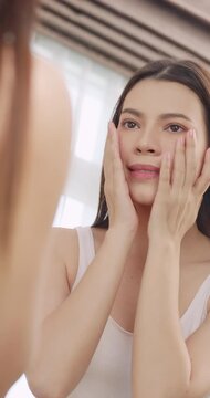 Vertical clip. Beautiful young woman applies moisturizer to her face while standing in front of a mirror.