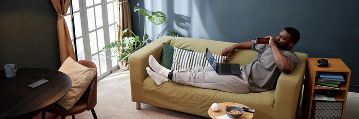 Website header shot of young adult Black man reclining on sofa drinking from bottle while using...