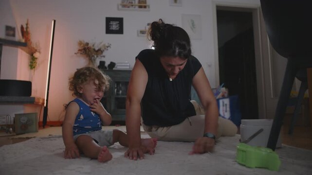 Mother rubbing and cleaning carpet to remove a stain while toddler cries beside her seeking attention, candid indoor family moment showing daily parenting challenges