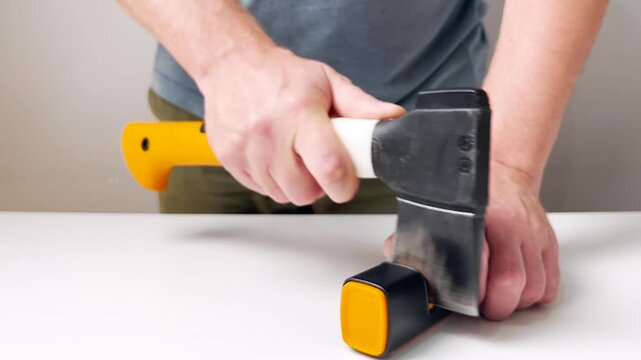 Close up video of a man sharpening an axe with a sharpening tool at home. Handheld process, metal blade maintenance, preparation for work, safety, household tasks, tool care
