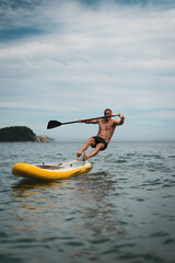 Athletic man loses balance and falls from paddle board into sea during summer fun. Action moment full of energy and laughter on warm vacation day with ocean adventure vibes.