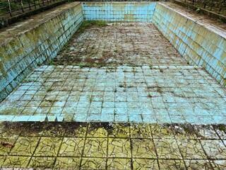 Abandoned, overgrown, and decaying swimming pool, showing faded teal tiles and concrete.
