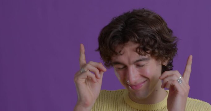 Close-up video clip of a smiling young adult male dancing with his fingers pointing and eyes closed against a purple studio background. He has dark curly hair and is wearing casual clothing.