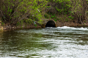 Naklejka premium Flowing river water near a concrete drainage pipe surrounded by green foliage in a serene natural setting during the daytime