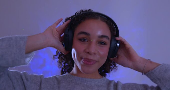Video clip of a young adult female smiling in the studio while listening to music through wireless headphones. She has brunette curly hair and she is illuminated by blue neon lights.